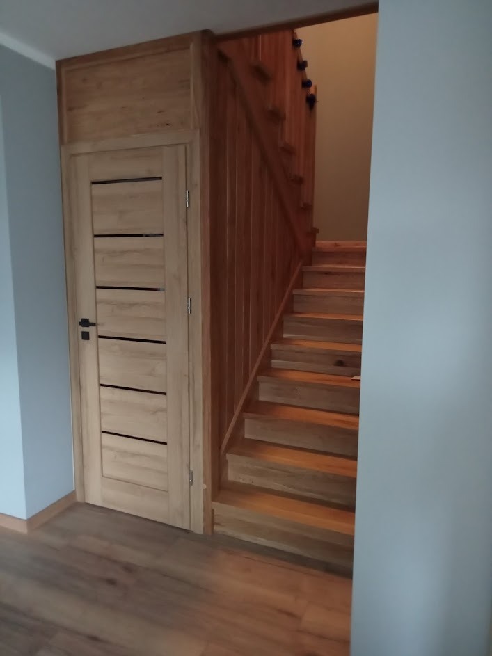 Drewniane schody z balustradą i drzwi z poziomymi przeszkleniami w jasnym odcieniu drewna, widoczne z perspektywy korytarza.