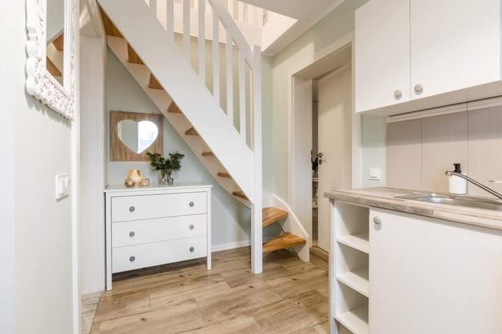 Jasne wnętrze z białymi schodami z drewnianymi stopniami, komodą z dekoracjami, lustrem w kształcie serca i fragmentem zabudowy kuchennej.