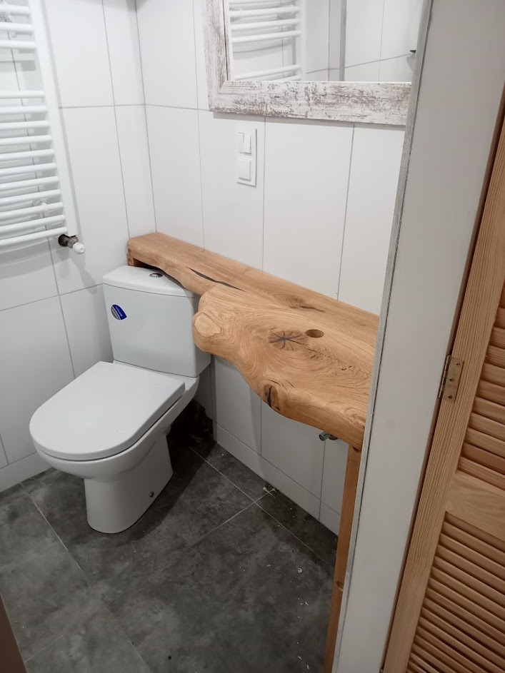 Blat łazienkowy z litego drewna o nieregularnym kształcie, zamontowany nad toaletą w nowoczesnej łazience z białymi płytkami i szarą podłogą.