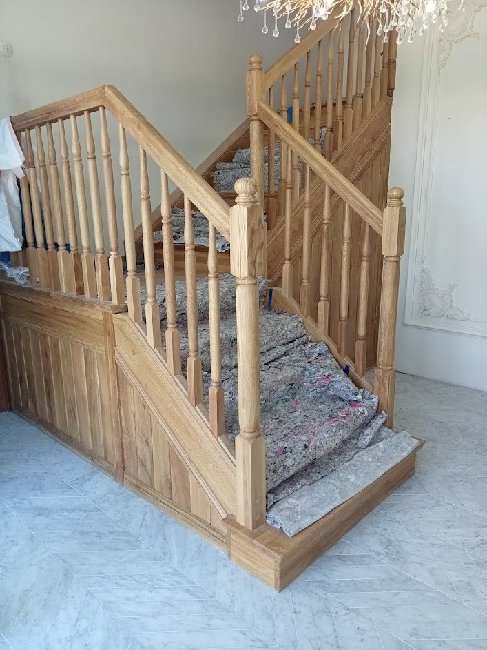 Drewniane schody z balustradą w trakcie montażu, zabezpieczone folią malarską, widoczne zdobienia na ścianie i żyrandol kryształowy.