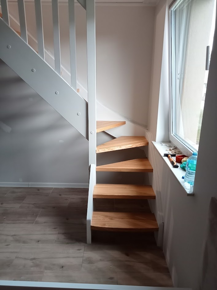 Drewniane schody z białymi barierkami prowadzące na antresolę, widok z dołu, na parapecie okiennym narzędzia i butelka z wodą.