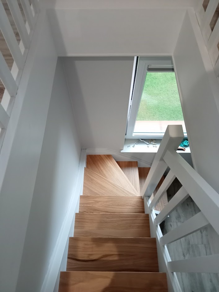 Drewniane schody z jasnego drewna, białe barierki, widok z góry na dół, okno dachowe z widokiem na zieleń.