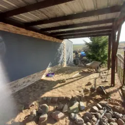 Budowa zadaszenia tarasu z elementami kamiennymi, widoczne narzędzia, taczka i częściowo wykończona elewacja z imitacji kamienia.