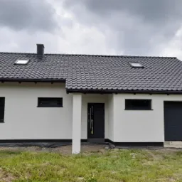 Nowoczesny dom jednorodzinny z białą elewacją, czarną stolarką okienną i grafitową dachówką, widok z zewnątrz na tle pochmurnego nieba.