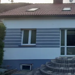 Wykończona elewacja domu jednorodzinnego z szarymi pasami, białymi oknami i schodami wejściowymi z kostki brukowej.