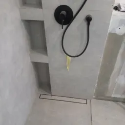 Nowoczesny prysznic walk-in z szarymi płytkami, czarną armaturą i odpływem liniowym, widoczne półki wnękowe w ścianie, fragment żółtej taśmy zabezpieczającej.