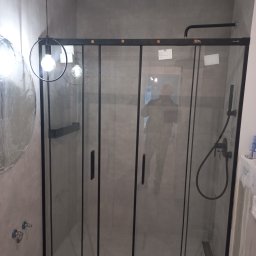 Otobud - Nowoczesna kabina prysznicowa z czarnymi ramami i szklanymi ścianami w łazience wykończonej szarymi płytkami. Widoczne elementy armatury, minimalistyczna lampa wisząca i odbicie osoby w szybie.