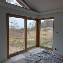 Otobud - Surowe wnętrze z dużym, trzyskrzydłowym oknem z drewnianą ramą, wychodzącym na krajobraz w Kamieniu Pomorskim, podłoga zabezpieczona folią malarską, ściany w odcieniach szarości przed wykończeniem.