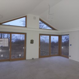 Otobud - Wnętrze w stanie deweloperskim z dużymi oknami i szarą podłogą; widoczne niedokończone prace wykończeniowe, wystające kable i zabezpieczenia na oknach.