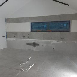 Otobud - Wnętrze w trakcie wykończenia: szare płytki podłogowe, ściany częściowo z szarymi płytkami, okno zabezpieczone folią, widoczne instalacje elektryczne i hydrauliczne, białe przewody na podłodze.