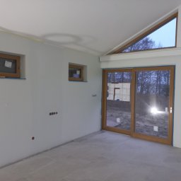 Otobud - Wnętrze w trakcie prac wykończeniowych: surowe ściany przygotowane do malowania, widoczne puszki elektryczne, drewniane okna z niebieską taśmą malarską, szare płytki podłogowe i duże przesuwne...