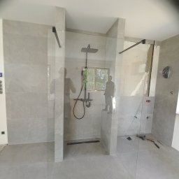 Otobud - Nowoczesna kabina prysznicowa walk-in z szarymi płytkami, czarną armaturą i widocznymi narzędziami na podłodze podczas prac wykończeniowych.