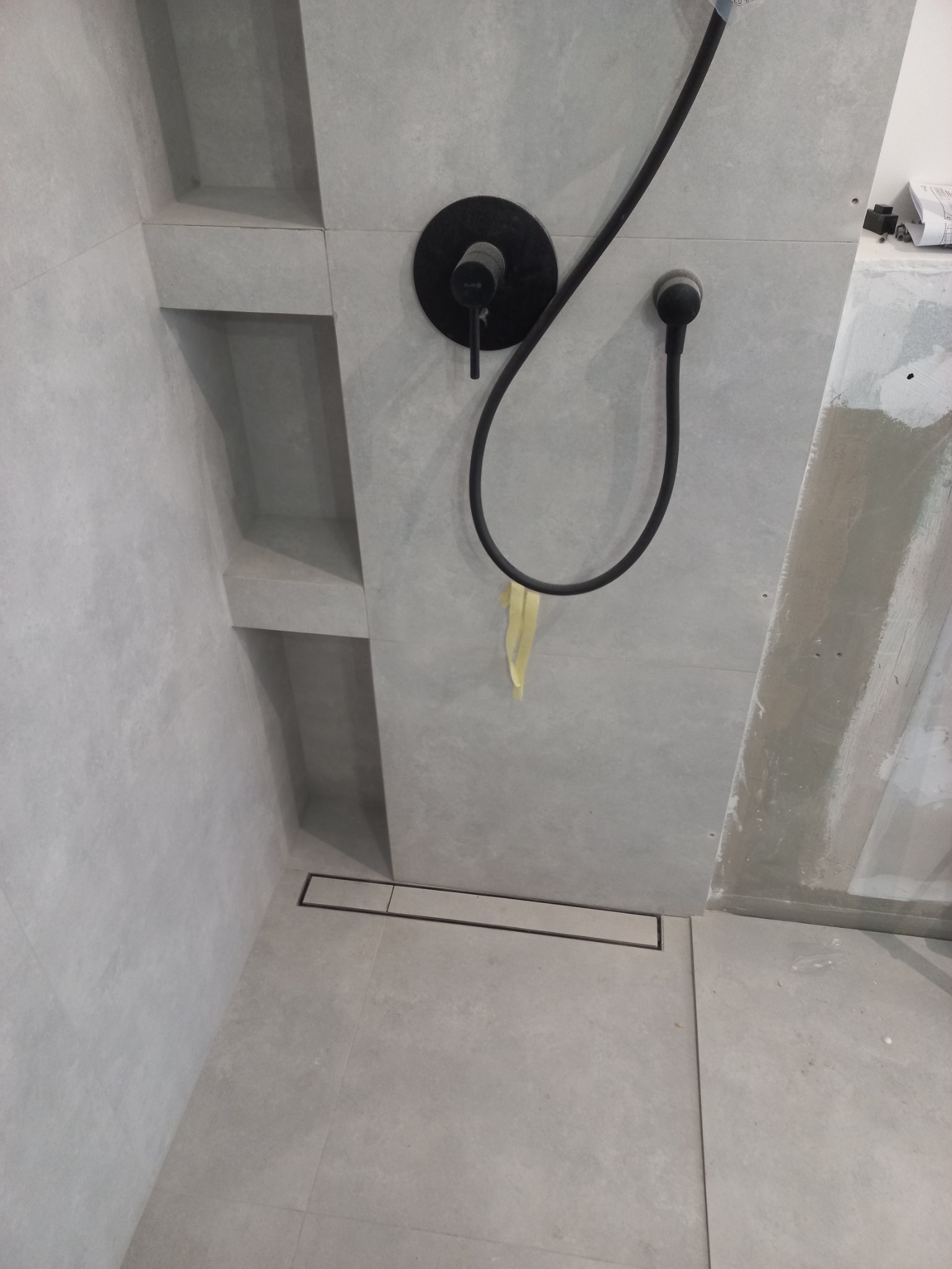 Nowoczesny prysznic walk-in z szarymi płytkami, czarną armaturą i odpływem liniowym, widoczne półki wnękowe w ścianie, fragment żółtej taśmy zabezpieczającej.
