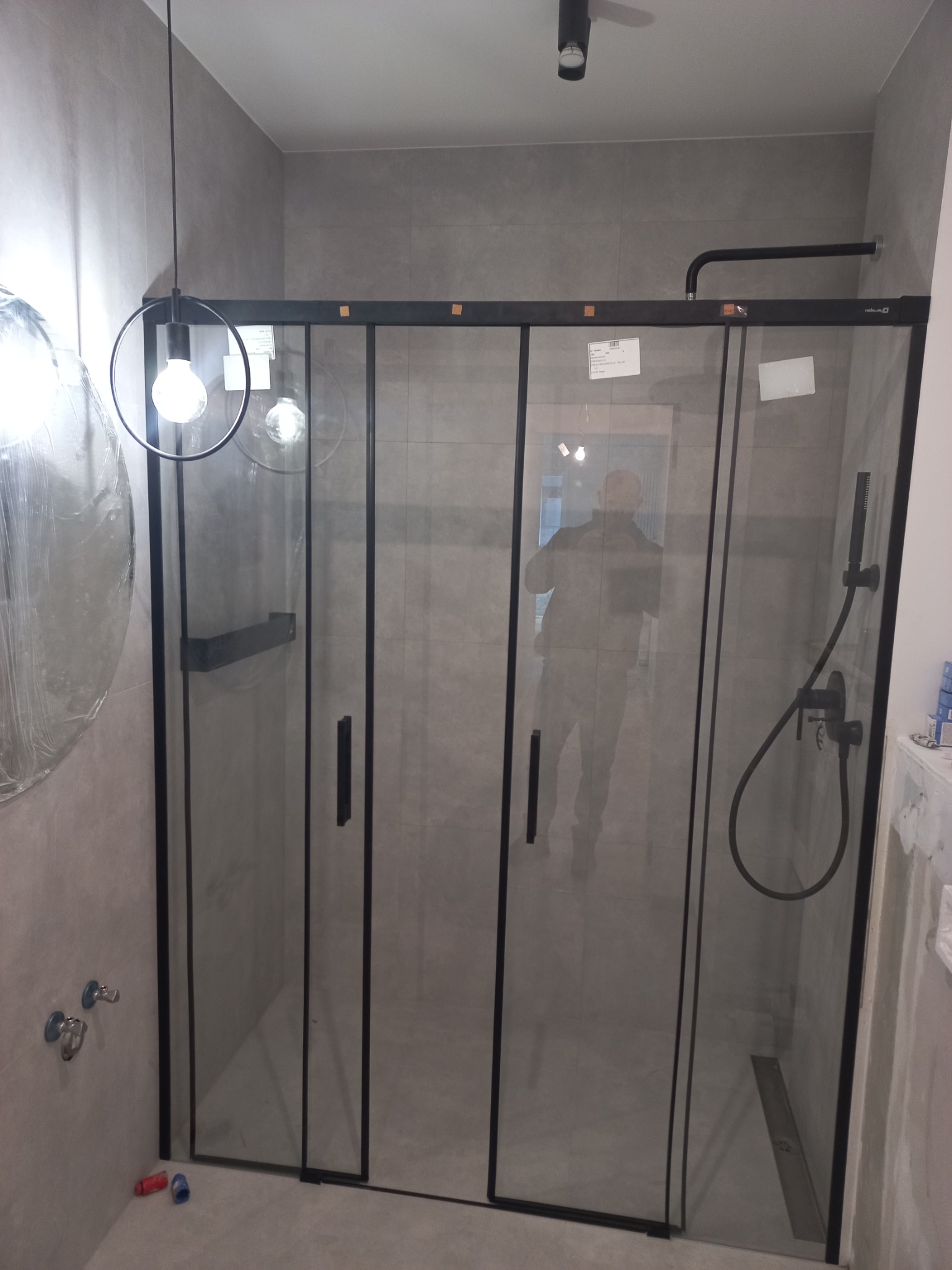 Nowoczesna kabina prysznicowa z czarnymi ramami i szklanymi ścianami w łazience wykończonej szarymi płytkami. Widoczne elementy armatury, minimalistyczna lampa wisząca i odbicie osoby w szybie.