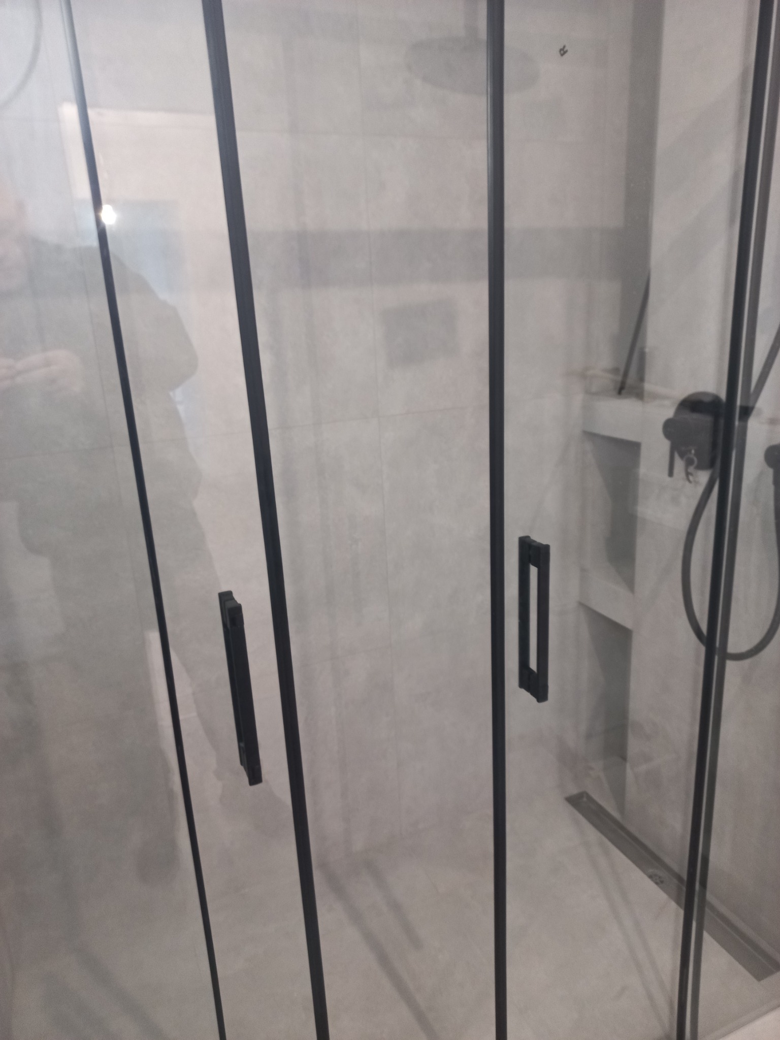 Szklana kabina prysznicowa z czarnymi profilami i armaturą, widoczny odpływ liniowy w posadzce, szare płytki na ścianach.