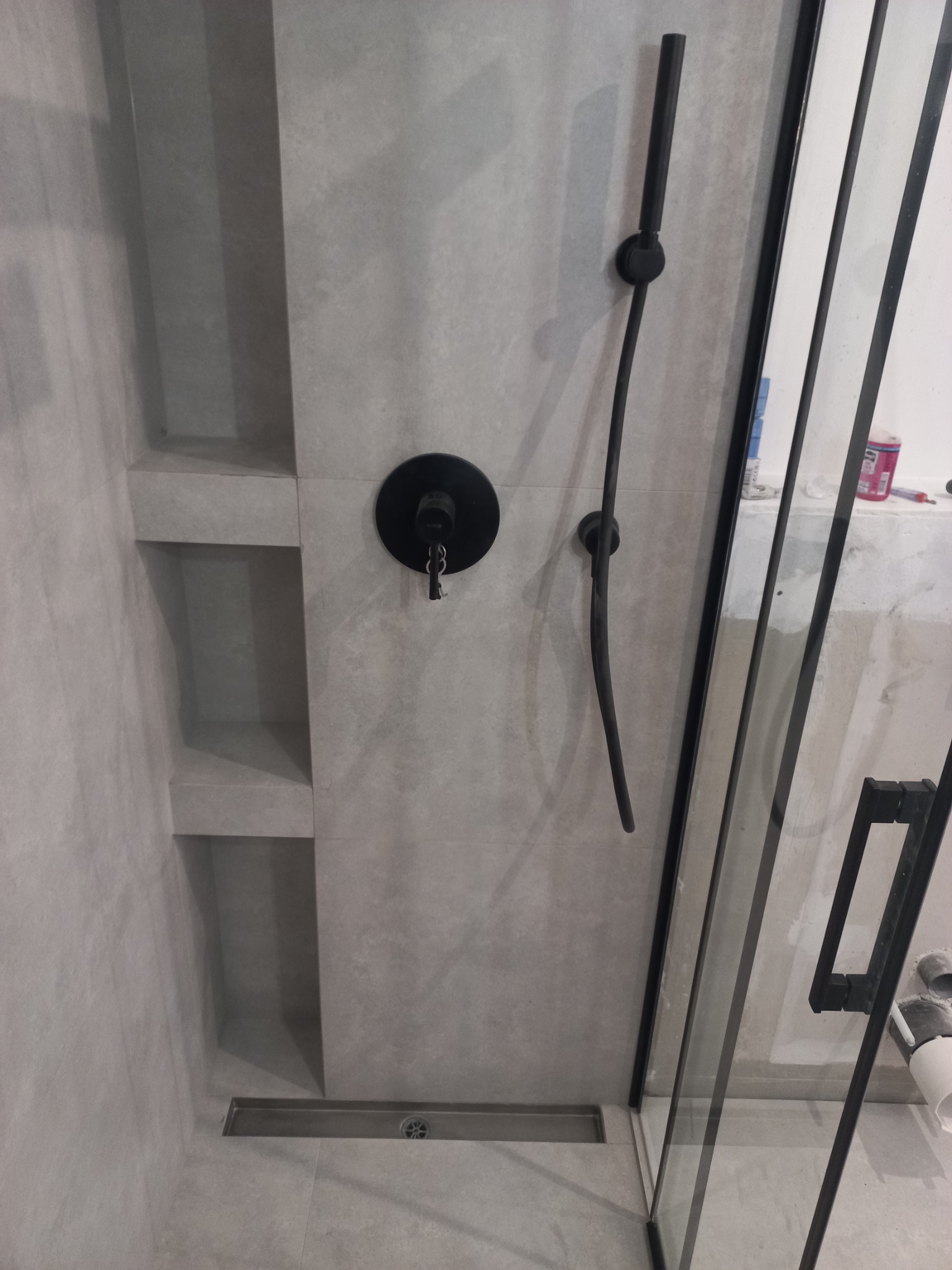 Szara łazienka z czarną armaturą: bateria prysznicowa z deszczownicą, wbudowane półki w ścianie, odpływ liniowy i szklana kabina prysznicowa z czarnymi profilami.