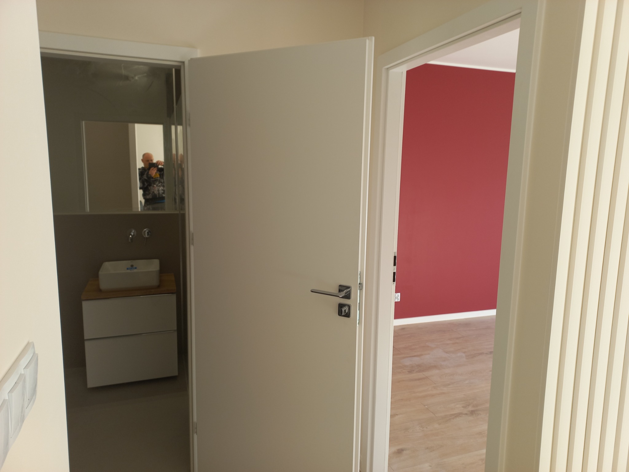 Otwarta, biała wewnętrzna drzwi, widok na wykończoną łazienkę z białą umywalką na szafce i odbicie fotografa w lustrze oraz pokój z czerwoną ścianą i drewnianą podłogą.