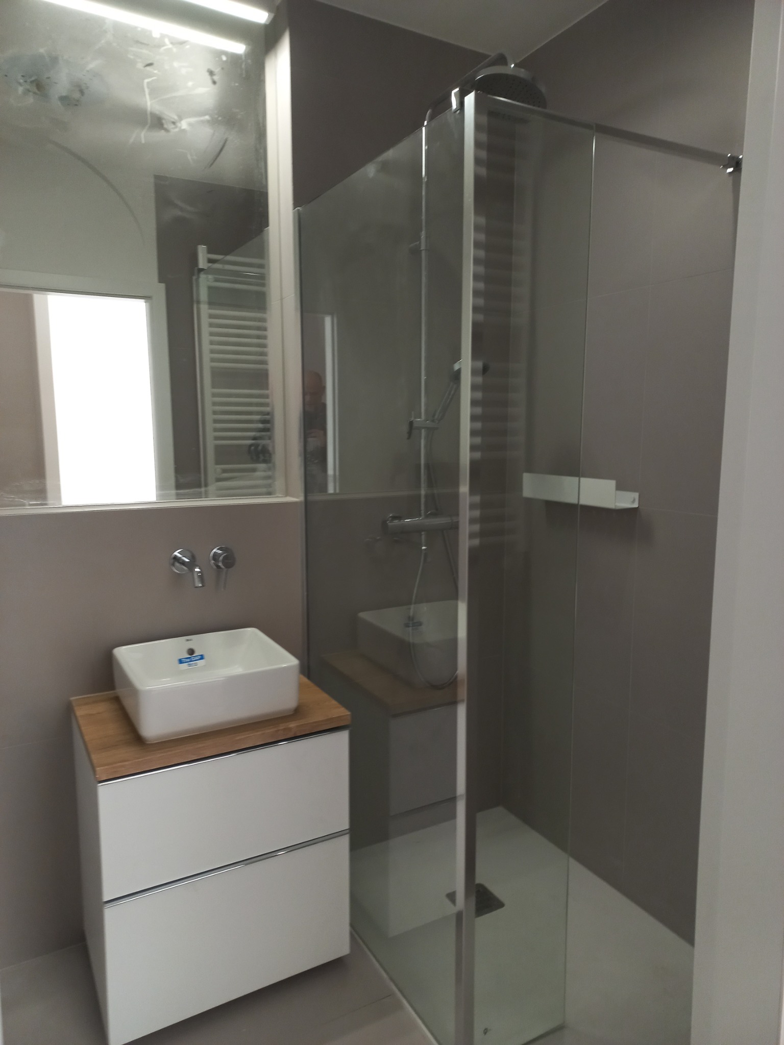 Nowoczesna łazienka z kabiną prysznicową ze szklanymi drzwiami i białą umywalką nablatową na szafce z drewnianym blatem, widoczne baterie ścienne i lustro z odbiciem grzejnika.