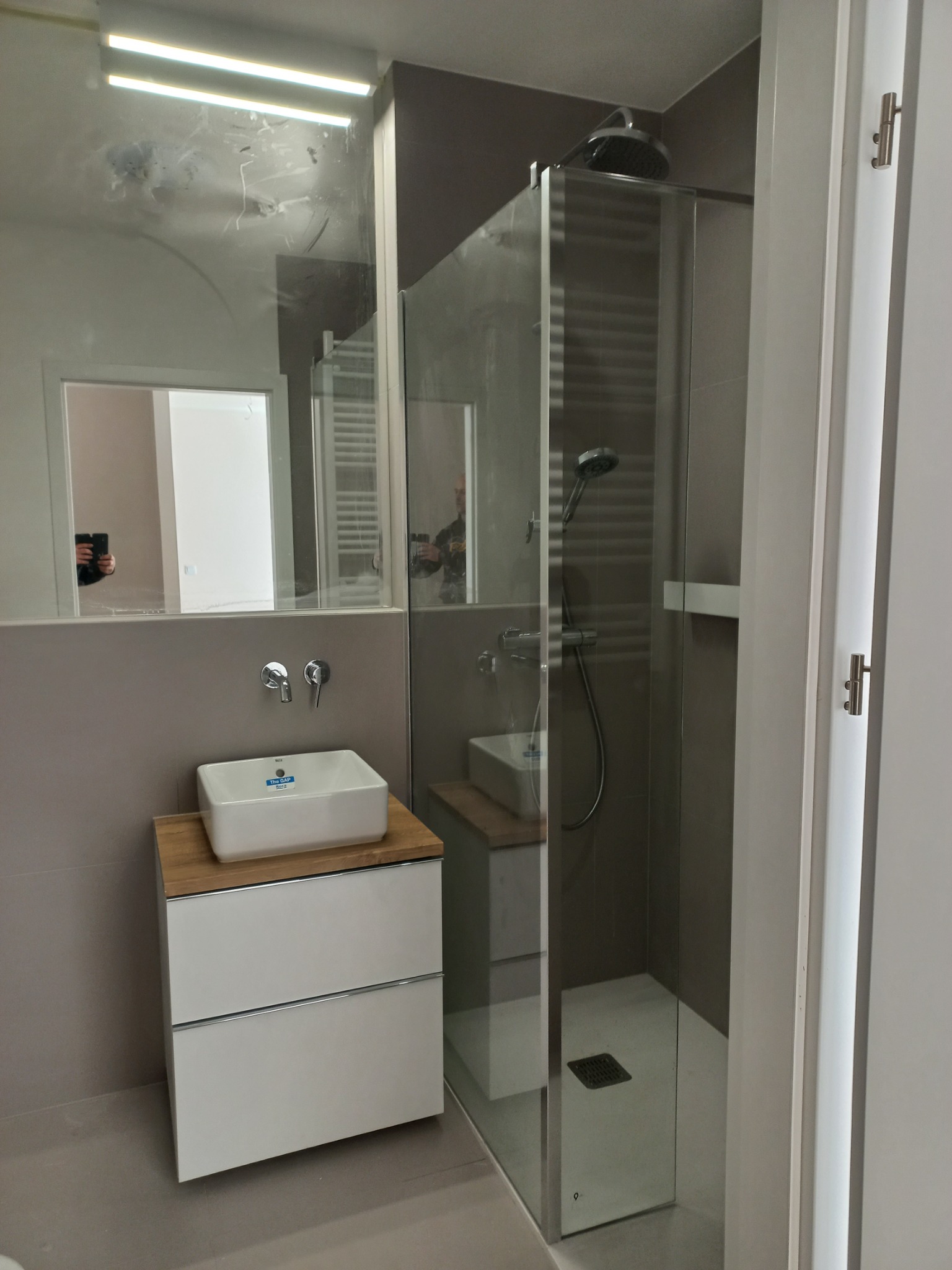 Nowoczesna łazienka po remoncie: kabina prysznicowa ze szklanymi drzwiami, biała umywalka nablatowa na szafce z drewnianym blatem, duże lustro z widocznymi smugami.