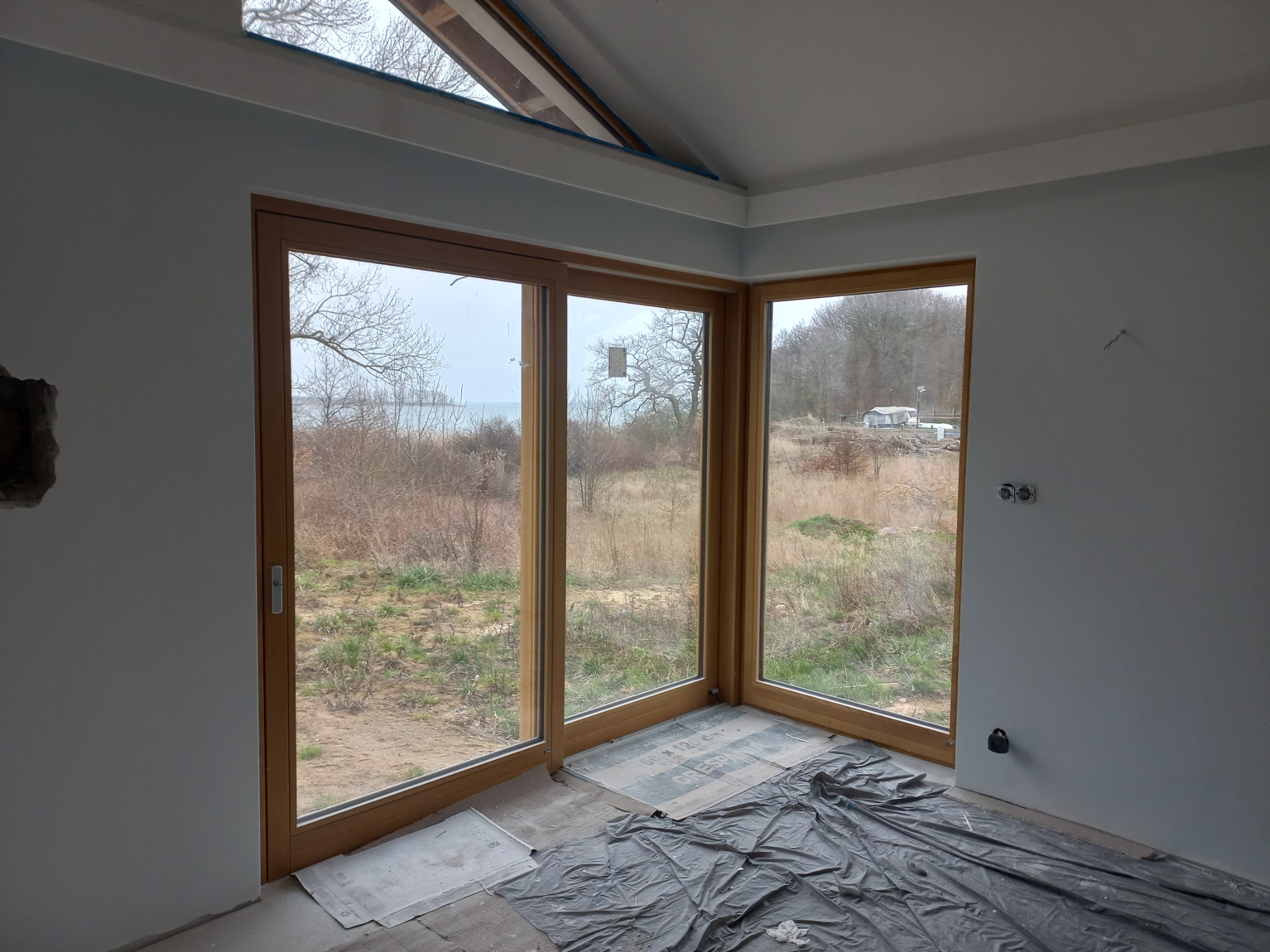 Surowe wnętrze z dużym, trzyskrzydłowym oknem z drewnianą ramą, wychodzącym na krajobraz w Kamieniu Pomorskim, podłoga zabezpieczona folią malarską, ściany w odcieniach szarości przed wykończeniem.