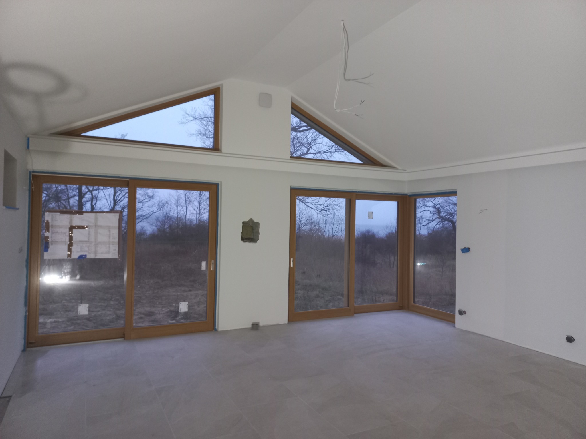 Wnętrze w stanie deweloperskim z dużymi oknami i szarą podłogą; widoczne niedokończone prace wykończeniowe, wystające kable i zabezpieczenia na oknach.