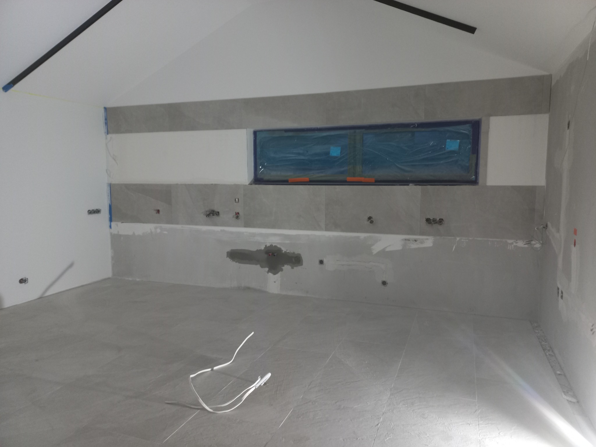 Wnętrze w trakcie wykończenia: szare płytki podłogowe, ściany częściowo z szarymi płytkami, okno zabezpieczone folią, widoczne instalacje elektryczne i hydrauliczne, białe przewody na podłodze.