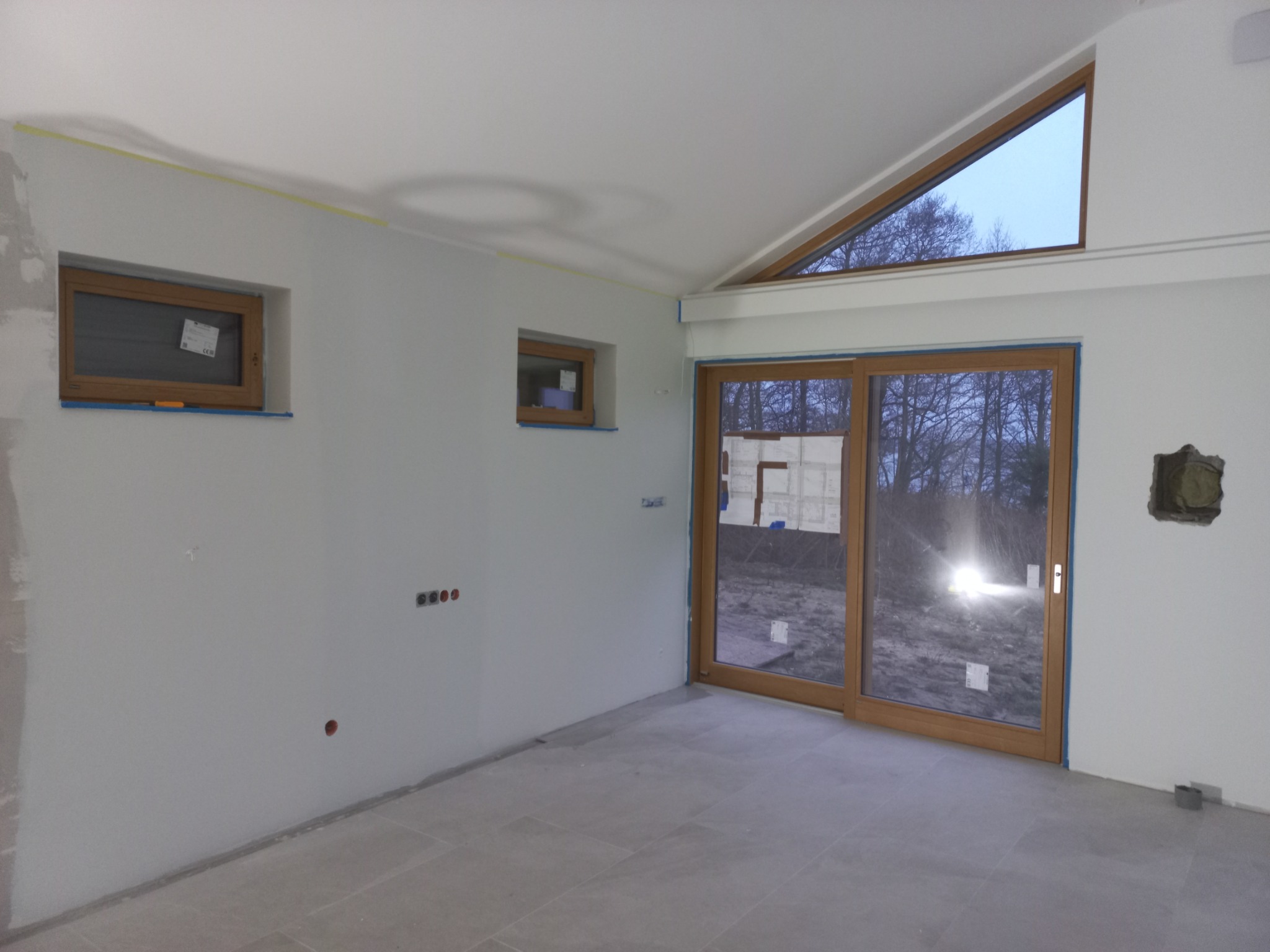 Wnętrze w trakcie prac wykończeniowych: surowe ściany przygotowane do malowania, widoczne puszki elektryczne, drewniane okna z niebieską taśmą malarską, szare płytki podłogowe i duże przesuwne...