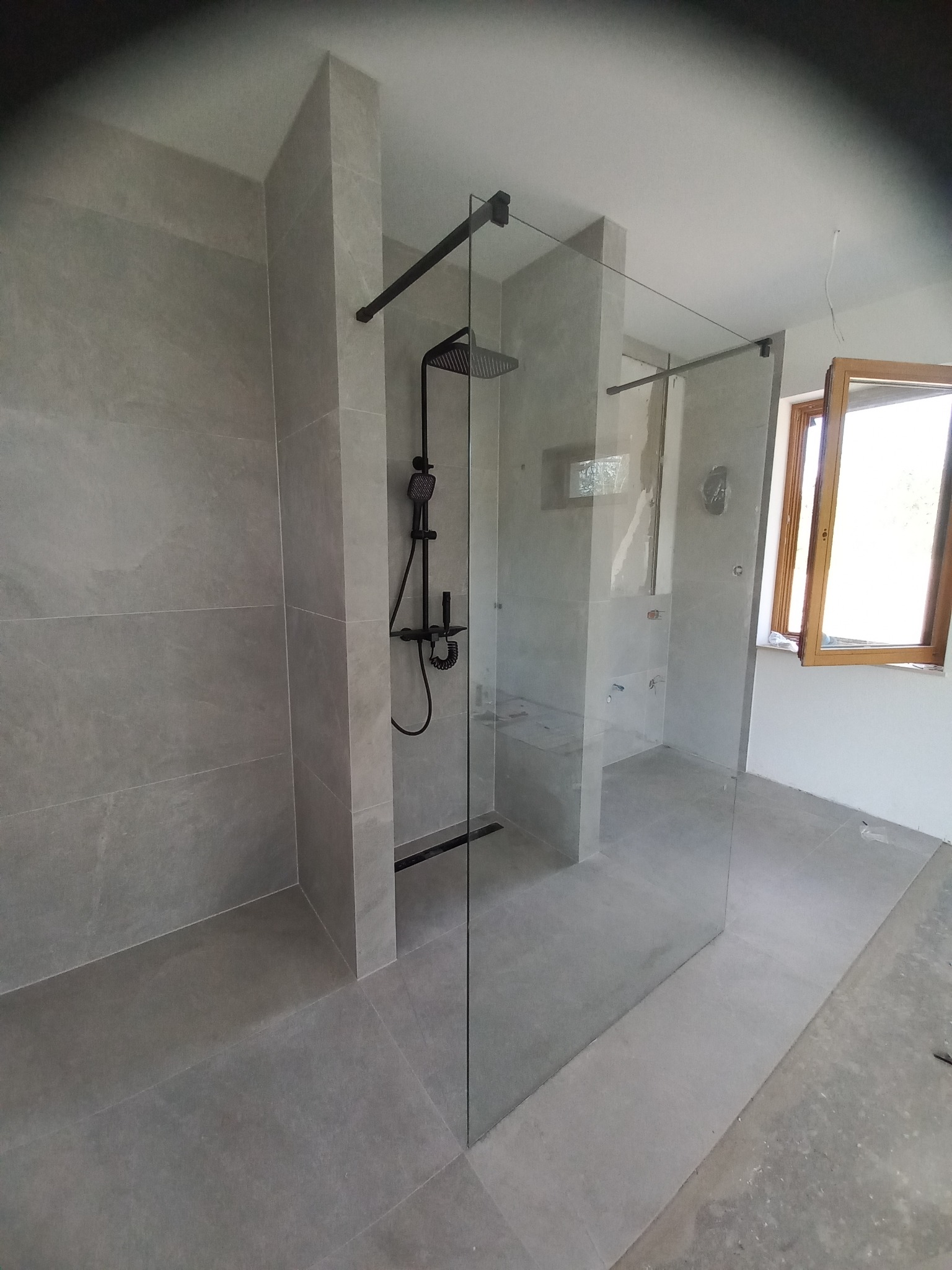 Nowoczesna kabina prysznicowa walk-in z czarną armaturą i szarymi płytkami, widok na okno w trakcie prac wykończeniowych.