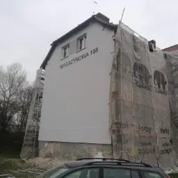 Malowanie elewacji budynku magazynowego z częściowo zdjętymi rusztowaniami i siatką ochronną z nadrukiem reklamowym farb Caparol, widoczny fragment samochodu na pierwszym planie.