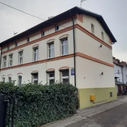 Odnowiona elewacja dwupiętrowego budynku w Inowrocławiu, z kremową fasadą i pomarańczowymi akcentami, widoczny numer budynku 104, barierka porośnięta bluszczem.