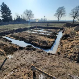 Wylane fundamenty domu jednorodzinnego na działce, widoczne szalunki i poziom betonu, w tle drzewa bez liści i błękitne niebo.