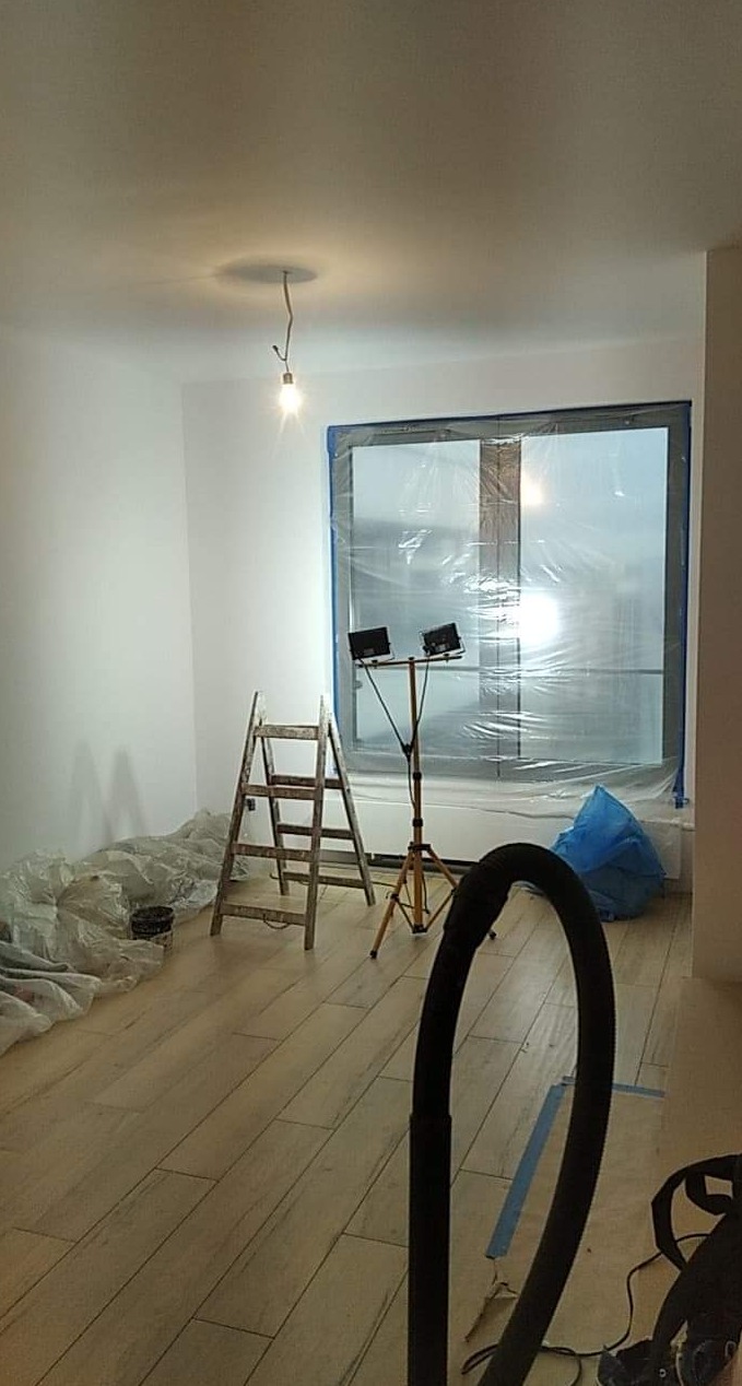 Przygotowanie pokoju do prac wykończeniowych: zabezpieczone okno folią, drabina, lampa oświetleniowa na statywie, podłoga wyłożona panelami, widoczny wąż odkurzacza.