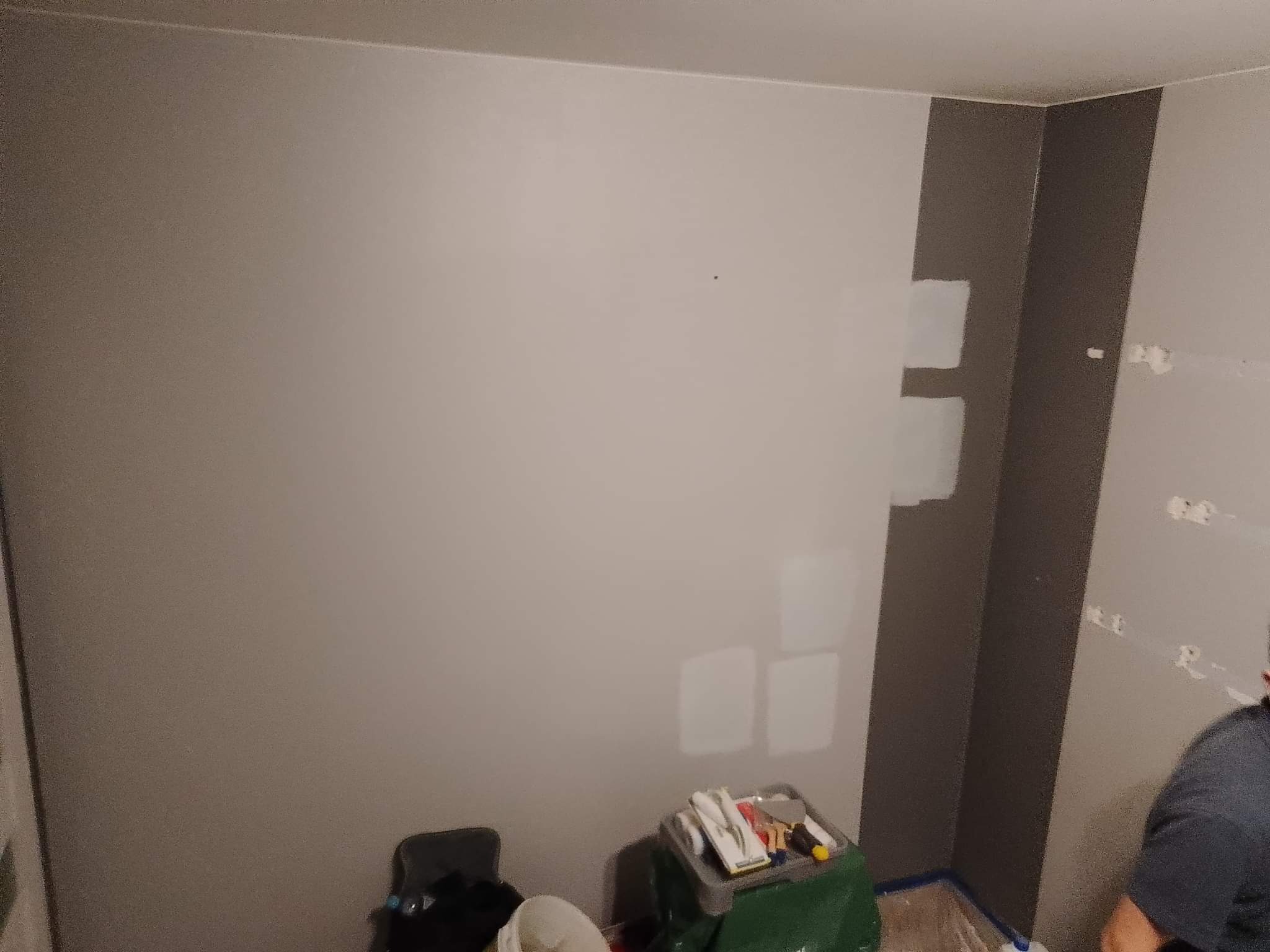 Ściana w trakcie malowania z widocznymi łatami świeżej farby, narzędzia malarskie na podłodze, ciemna pionowa linia na ścianie, prawdopodobnie fragment dekoracyjny.