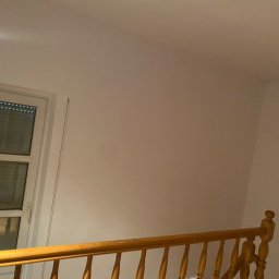 Adam kaczmarek - Biała ściana po odświeżeniu w Łodzi, widoczny fragment okna z żaluzjami i drewniana balustrada schodów.