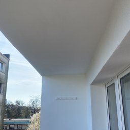 Adam kaczmarek - Odnowiony balkon z widokiem na sąsiedni budynek; świeżo pomalowane ściany i sufit, biała stolarka okienna, na ścianie zamontowany wieszak na ubrania.