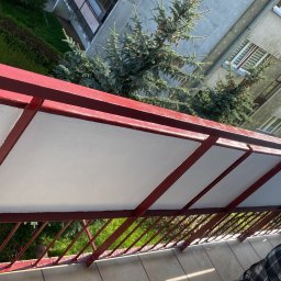 Adam kaczmarek - Widok z góry na odnowiony balkon z pomalowanymi na biało panelami i czerwoną metalową konstrukcją; w tle budynki mieszkalne i zieleń drzew.