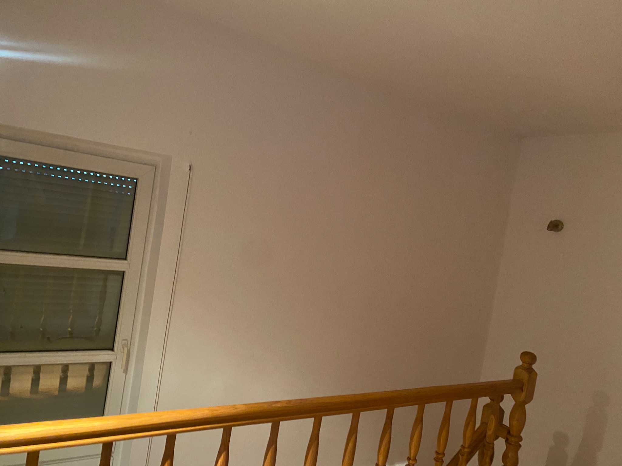 Biała ściana po odświeżeniu w Łodzi, widoczny fragment okna z żaluzjami i drewniana balustrada schodów.