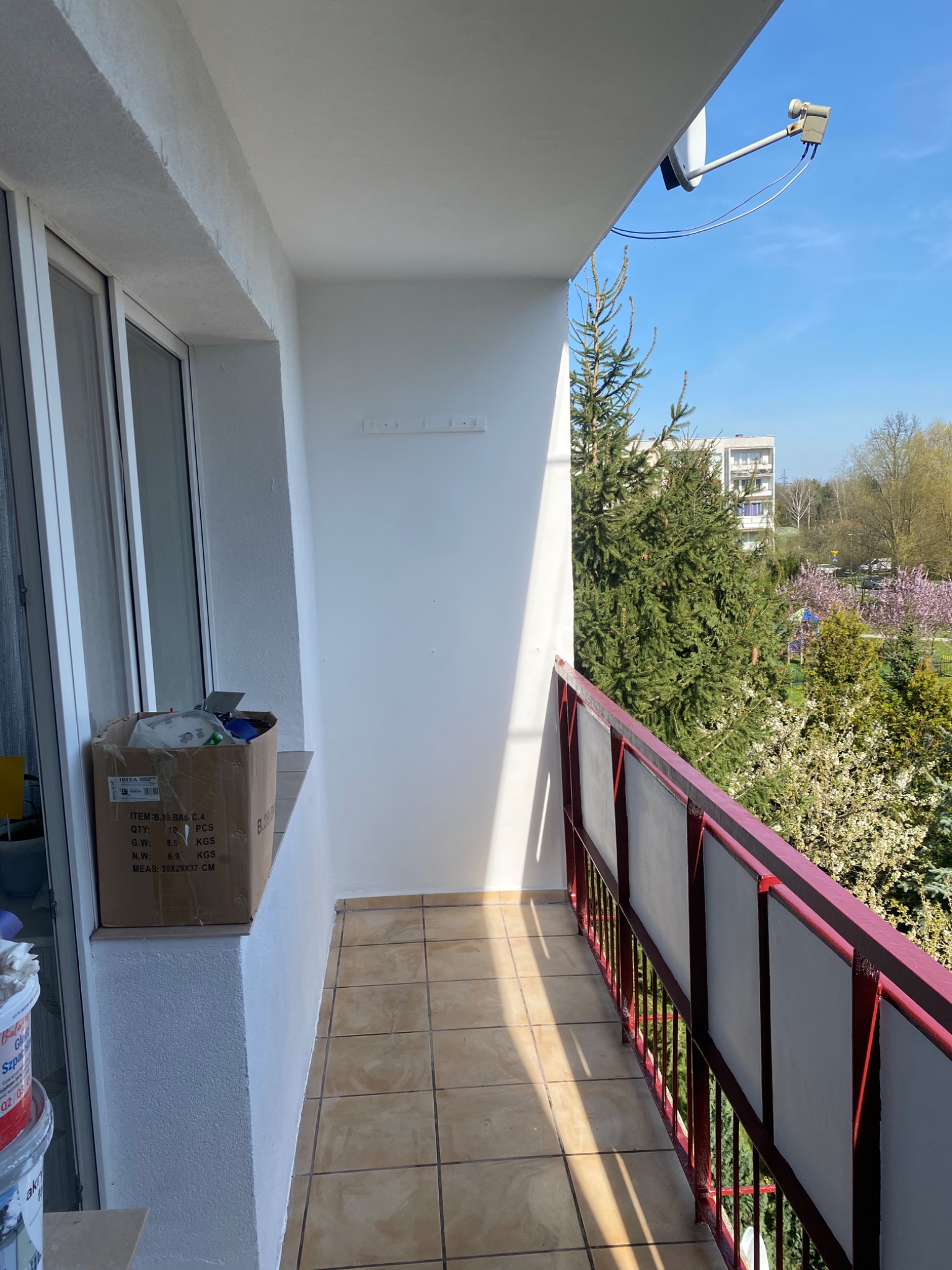 Białe, wyszpachlowane ściany balkonu z brązową terakotą na podłodze, widok na zieleń drzew i błękitne niebo, karton z akcesoriami i puszki z farbą w rogu balkonu.