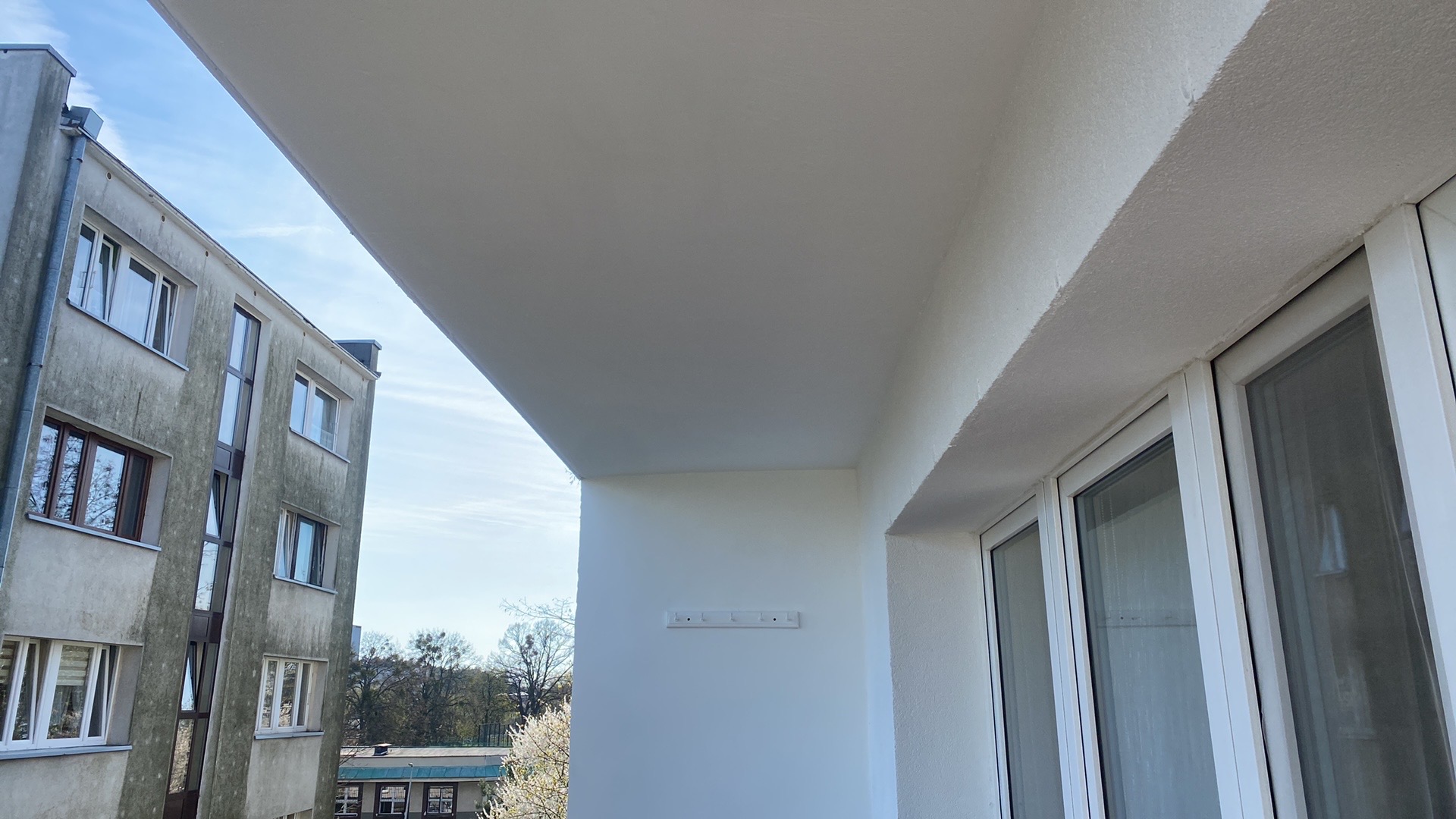 Odnowiony balkon z widokiem na sąsiedni budynek; świeżo pomalowane ściany i sufit, biała stolarka okienna, na ścianie zamontowany wieszak na ubrania.