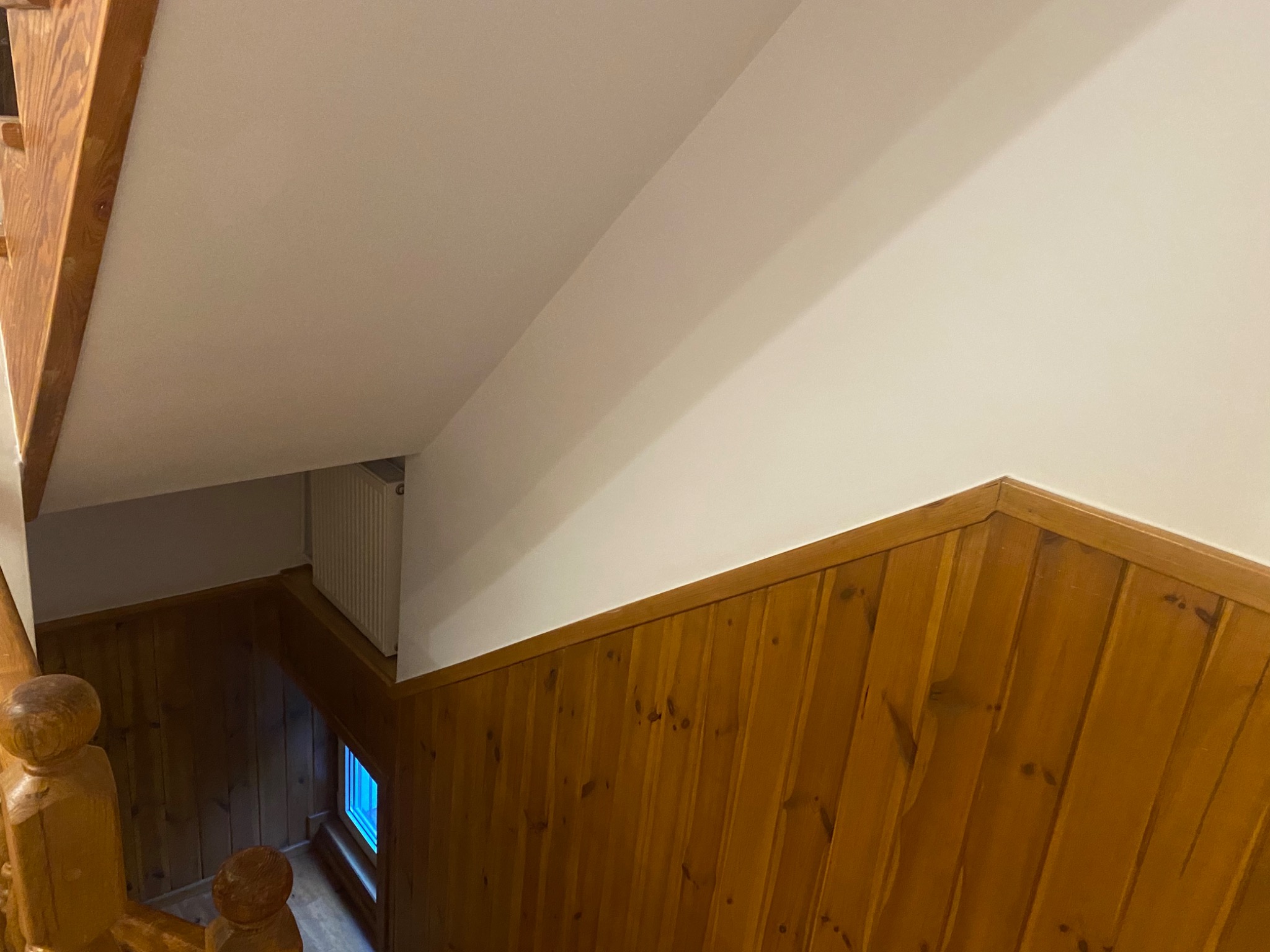 Wnętrze klatki schodowej z drewnianą boazerią do połowy wysokości ściany i świeżo pomalowaną, jasną górną częścią ściany oraz sufitem.