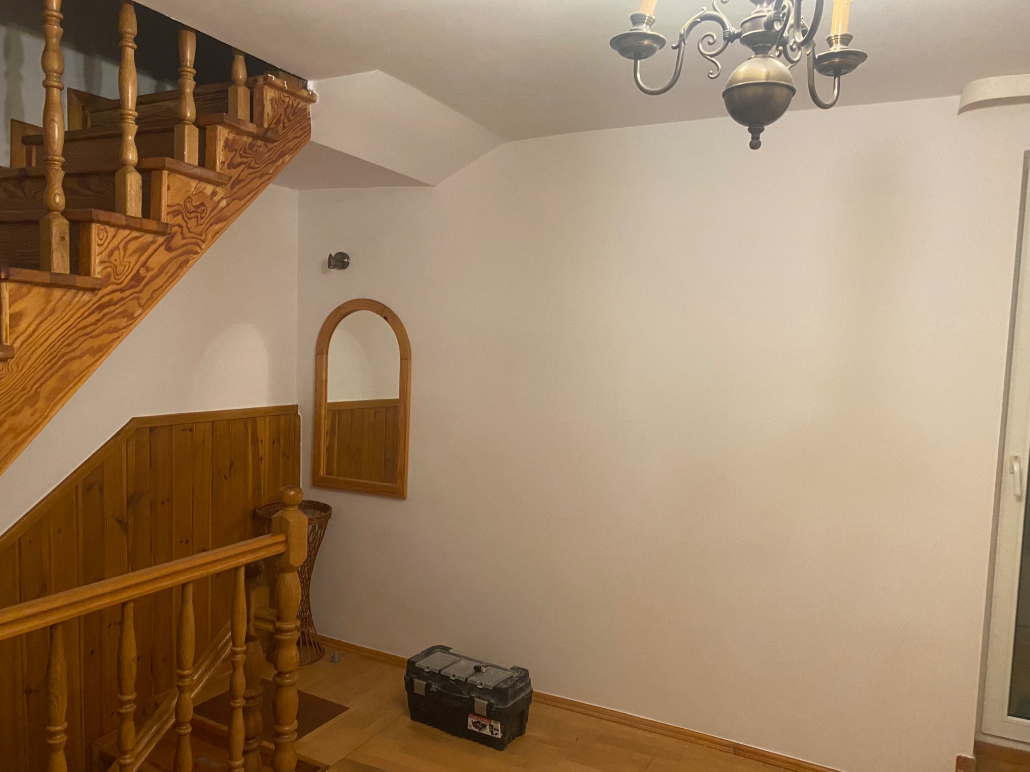 Wnętrze domu przed malowaniem: drewniane schody i boazeria, białe ściany, czarna skrzynka z narzędziami na podłodze, lustro w drewnianej ramie i metalowy żyrandol.