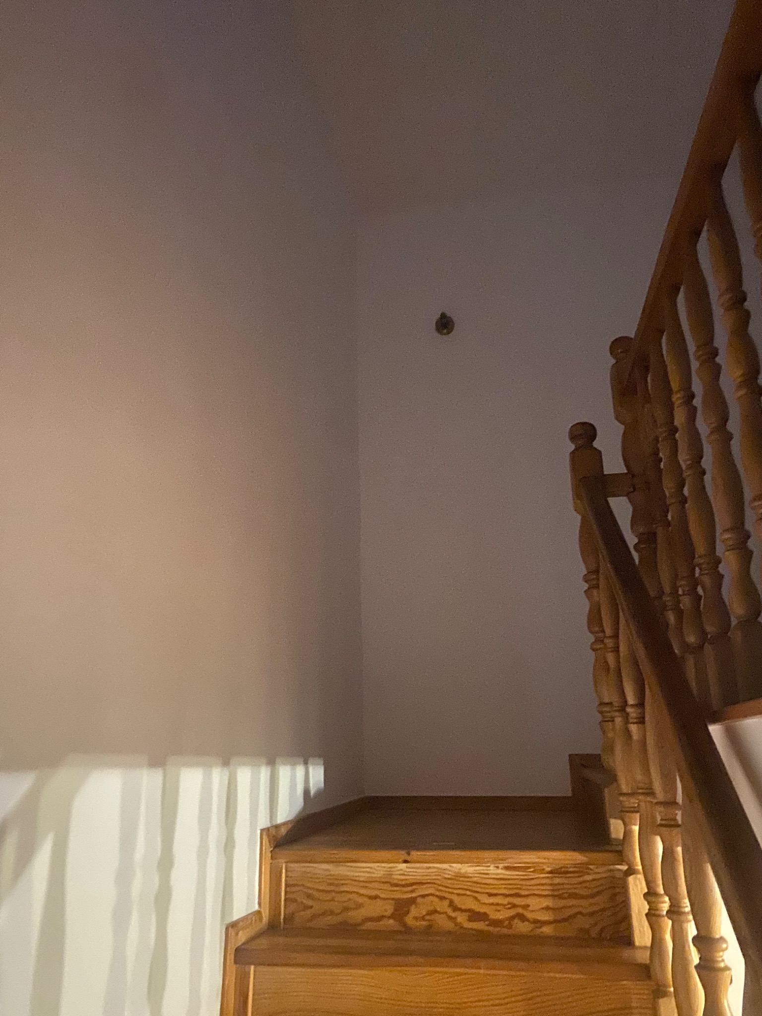 Fragment klatki schodowej z drewnianymi schodami i balustradą. Ściana pomalowana na jasny kolor z widocznym pojedynczym otworem po kołku. Na ścianie widoczne cienie rzucane przez balustradę.