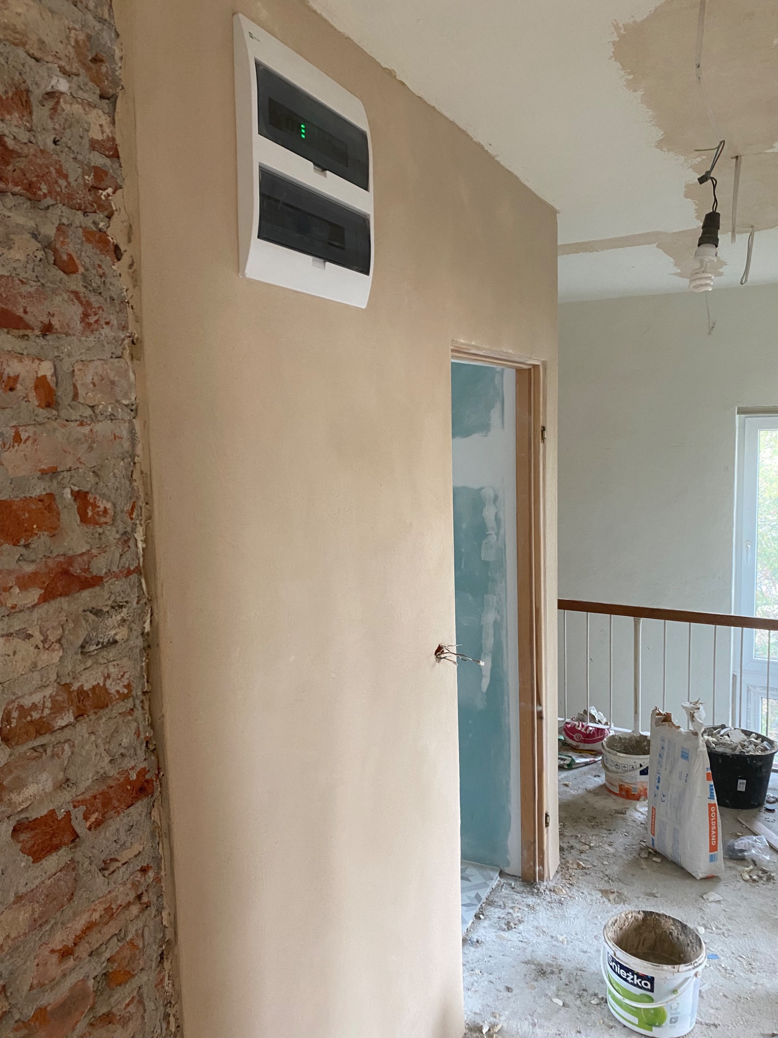 Wnętrze w trakcie prac wykończeniowych: fragment ściany z nałożoną warstwą gładzi, obok odsłonięta cegła, biała skrzynka elektryczna, widoczne worki z zaprawą i wiaderka po farbach, surowy stan...