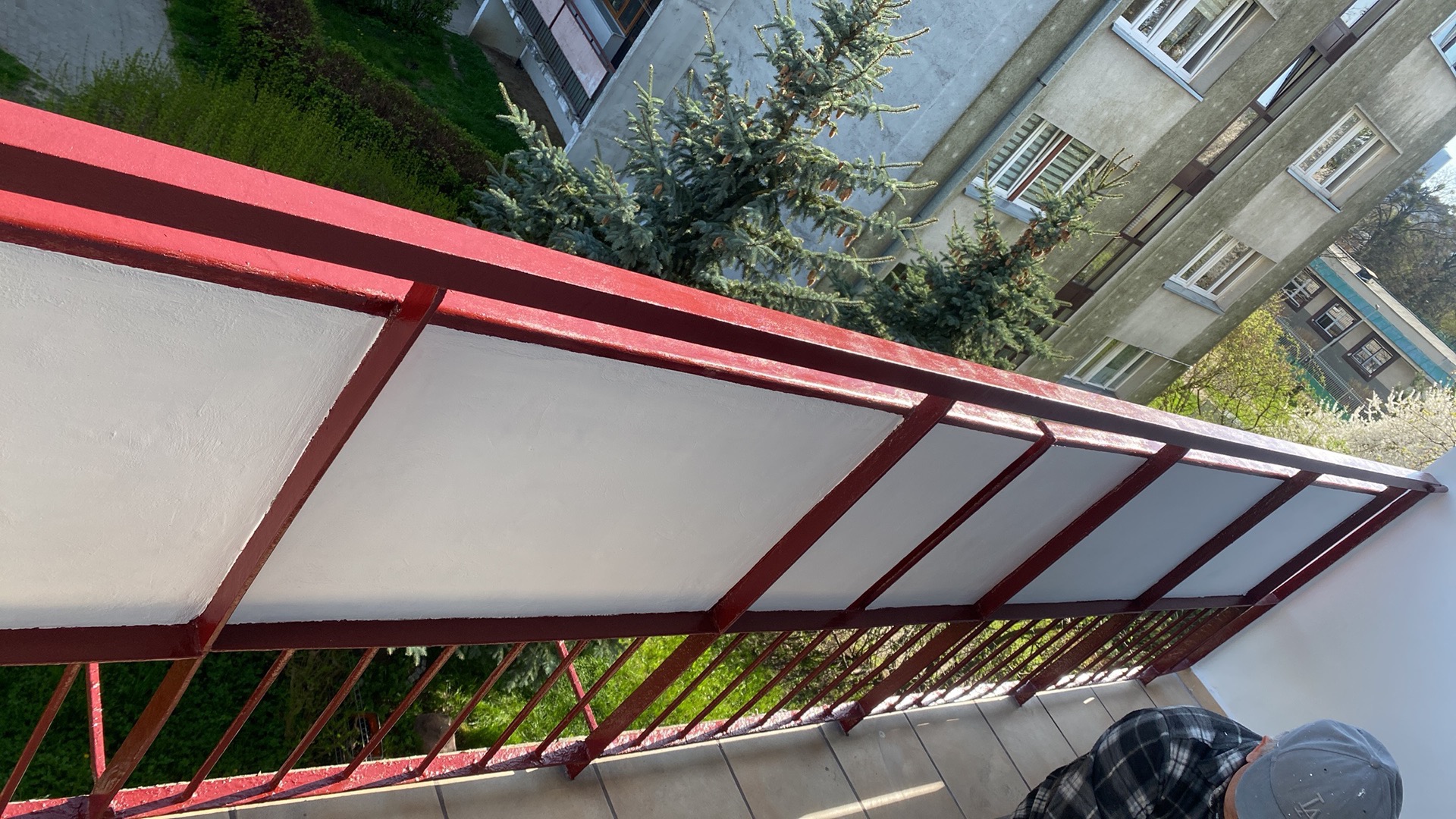 Widok z góry na odnowiony balkon z pomalowanymi na biało panelami i czerwoną metalową konstrukcją; w tle budynki mieszkalne i zieleń drzew.