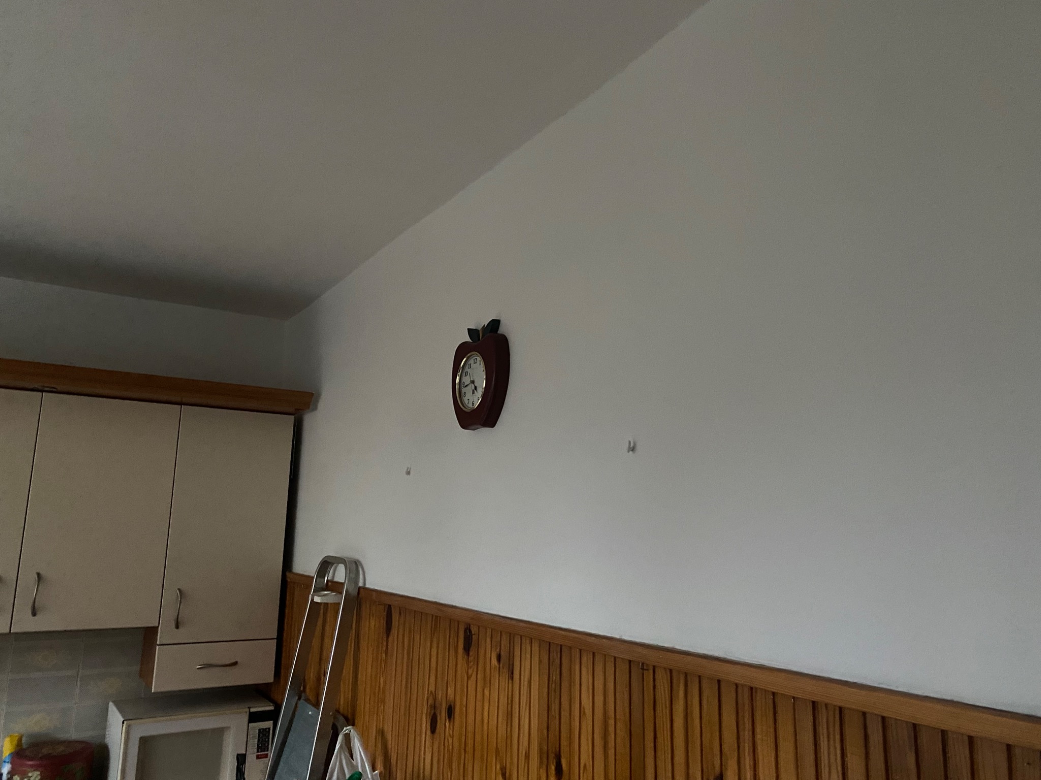 Ściana w kuchni po szpachlowaniu, z widocznymi śladami po kołkach, przygotowana do dalszych prac wykończeniowych, z drewnianą boazerią w dolnej części i zegarem w kształcie jabłka.