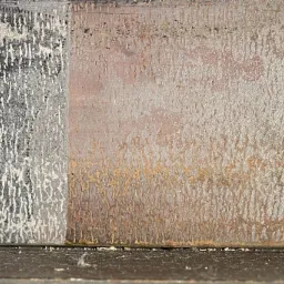 Metalowa powierzchnia podzielona na dwie części: lewa pokryta grubą warstwą białej farby, prawa z widoczną rdzawą strukturą, ukazująca efekt czyszczenia.