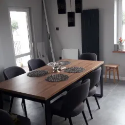 Nowoczesna jadalnia z drewnianym stołem na metalowych nogach, szare krzesła z tapicerowanym siedziskiem, szare płytki podłogowe, wiszące lampy z ażurowymi kloszami, grzejnik panelowy w kolorze...