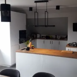 Nowoczesna kuchnia z białą zabudową, drewnianym blatem i czarnymi lampami wiszącymi o ażurowych kloszach oraz prostokątną lampą nad wyspą kuchenną.