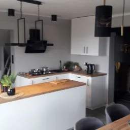 Nowoczesna kuchnia z białymi szafkami, drewnianym blatem wyspy i czarnymi lampami wiszącymi o ażurowych kloszach, widoczne przesuwne drzwi z czarną ramą i mlecznym szkłem.