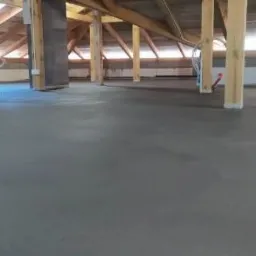 Świeżo wylana, gładka posadzka na poddaszu z drewnianą konstrukcją więźby dachowej i kolumnami wspierającymi.